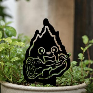 May include: Black metal garden stake featuring a whimsical design of a mountain with a face and a log. The stake is placed in a pot with green plants. The design has a cut-out style.