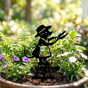 May include: A black metal garden stake silhouette of a mouse wearing a hat and apron, standing on a small ladder while holding a pair of garden shears. The mouse figure is positioned upright in the centre of a round planter filled with soil.