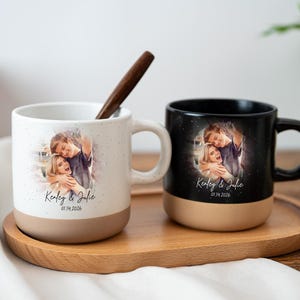 Puede incluir: Dos tazas de cerámica con impresiones fotográficas personalizadas. Una taza es blanca con un diseño moteado, la otra es negra. Ambas tazas tienen una base beige y un asa. Las tazas están sobre una bandeja de madera. Las tazas tienen el texto "Kenley & Julie" y una fecha.