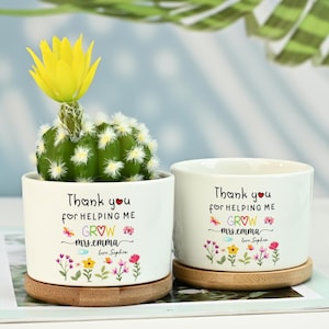 Könnte beinhalten: Zwei weiße Keramik-Pflanztöpfe mit Bambus-Untersetzern, jeweils mit einem kleinen Kaktus mit gelber Blüte. Die Töpfe tragen den Text "Thank you for helping me GROW mrs.emma love Sophia" und Blumendesigns.