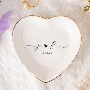 Könnte beinhalten: Eine weiße herzförmige Keramik-Ringschale mit goldenem Rand. Die Schale trägt die Initialen "J ♥ B" und das Datum "03.18.26" in schwarzer Schrift. Ein goldener Ring mit einer Perle liegt in der Nähe, zusammen mit einer Perlenkette und Blumen.