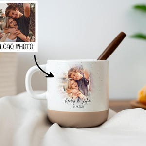 Puede incluir: Taza de cerámica blanca con base marrón claro, con una foto personalizada y el texto "Kenley & Julie 01.14.2026". Una cuchara de madera descansa dentro de la taza. La imagen también incluye una pequeña foto con el texto "UPLOAD PHOTO".