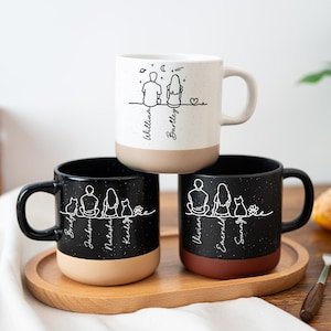 Puede incluir: Tres tazas de cerámica con diseños moteados en blanco y negro. Cada taza presenta un dibujo lineal de una pareja o familia con nombres debajo. Las tazas tienen un diseño de dos tonos con una base beige o marrón y asas negras.