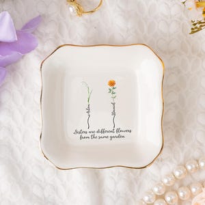 Puede incluir: Un cuenco de cerámica blanca con borde dorado, con ilustraciones florales y el texto "Sisters are different flowers from the same garden." El cuenco tiene los nombres "Arlen" y "Daniel".