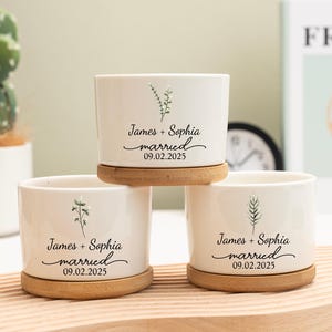 以下が含まれることがあります： 竹の受け皿が付いた、白いセラミック製の植木鉢3つ。各植木鉢には植物のイラストと、黒い筆記体で「James + Sophia married 09.02.2025」と書かれています。背景にはサボテンが見えます。