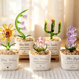 May include: A collection of glass flower sculptures in small white pots. The flowers include a sunflower, a lily, a cactus, a rose, and a lavender. Each pot has a message for a teacher. The pots are filled with small pebbles.