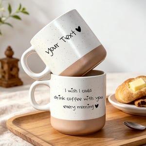 Può includere: Due tazze in ceramica impilate con una sezione superiore bianca maculata e una base color marrone chiaro. La tazza superiore presenta il testo "Your Text" con un'icona a forma di cuore. La tazza inferiore presenta il testo "I wish I could drink coffee with you every morning" con un cuore.
