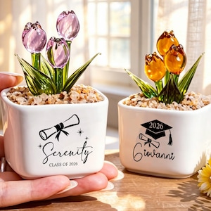 May include: Two small, square white ceramic pots with glass flower arrangements. One has purple tulips, the other orange. Each pot features a graduation design with a name and the year 2026.