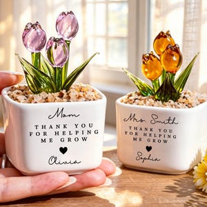 Pode incluir: Duas pequenas floreiras quadradas brancas com flores de tulipa de vidro. Uma floreira diz "Mãe, OBRIGADO POR ME AJUDAR A CRESCER, Olivia" e a outra diz "Sra. Smith, OBRIGADO POR ME AJUDAR A CRESCER, Sophia".