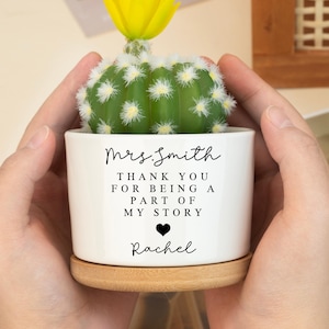 May include: A small, white ceramic pot with a cactus and a yellow flower. The pot is inscribed with "Mrs. Smith, THANK YOU FOR BEING A PART OF MY STORY, Rachel". It rests on a small, round wooden coaster. The pot is held by hands.