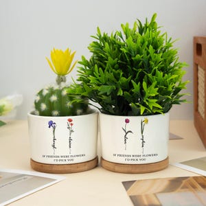 Könnte beinhalten: Zwei weiße Keramik-Blumentöpfe mit Holzsockeln. Jeder Topf hat ein anderes Blumenmuster und einen personalisierten Namen, der vertikal am Stiel der Blume entlang geschrieben ist. Der Text auf den Töpfen lautet "Wenn Freunde Blumen wären, würde ich dich wählen".
