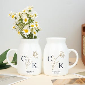 May include: Two white ceramic mugs with handles, featuring a gold floral design and the initials "C" and "K". The mugs have the text "Engaged 09.08.24" printed on them. A white ceramic vase with a handle is filled with white daisies.