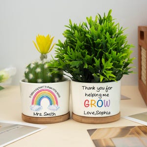 May include: Two white ceramic pots with plants, one with a cactus and the other with a green plant. The first pot has a rainbow design and the text "It takes a big heart to shape little minds" and "Mrs. Smith". The second pot has the text "Thank you for helping me GROW Love, Sophia".