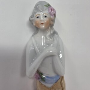May include: A porcelain figurine of a woman with grey hair and a pink flower. She wears a white top, a blue sash, and a tan skirt. The figurine has a delicate, hand-painted face and is likely a vintage collectible.
