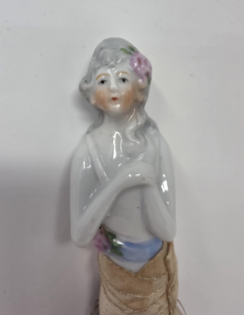 May include: A porcelain figurine of a woman with grey hair and a pink flower. She wears a white top, a blue sash, and a tan skirt. The figurine has a delicate, hand-painted face and is likely a vintage collectible.