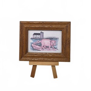 May include: A small rectangular picture frame with a wood grain finish, resting on a light wood easel. The print inside features a watercolor illustration of a large pink pig followed by several piglets in front of a farm building and silo.