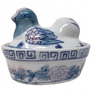 May include: A white ceramic lidded dish featuring a blue and pink hand-painted duck-shaped lid. The base of the bowl is decorated with a blue geometric Greek key pattern along the rim and a floral vine motif around the lower exterior.