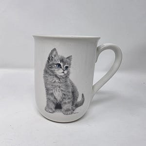 May include: White ceramic mug featuring a detailed illustration of a gray kitten with blue eyes. The kitten is sitting and looking to the side. The mug has a curved handle and a slightly flared rim.