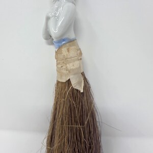May include: A vintage doll-shaped brush with a porcelain head and arms. The doll has a painted face, a grey hairstyle with pink flowers, and a white top. The brush portion is made of brown bristles, and the doll's waist is wrapped with a beige ribbon.
