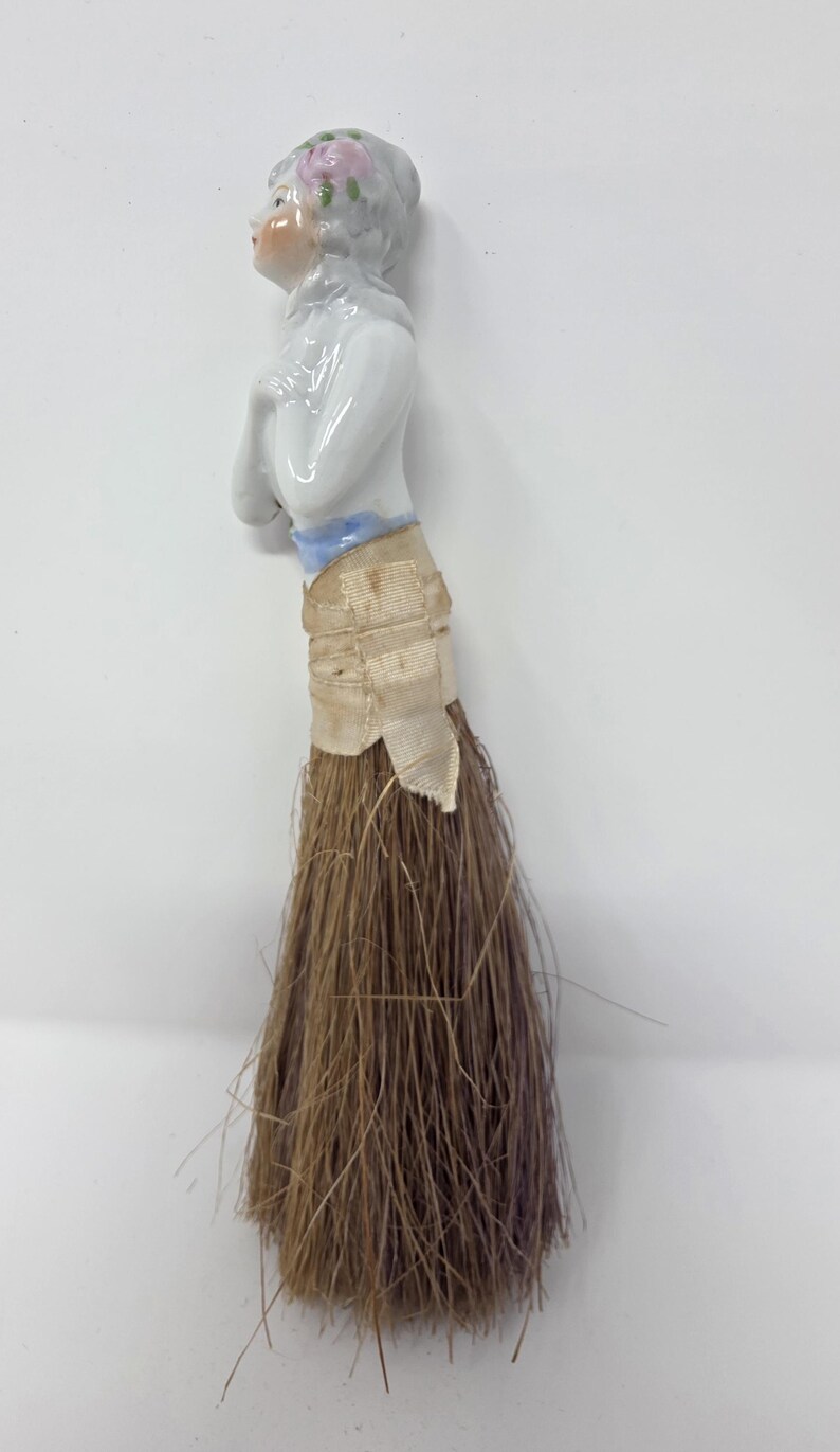 May include: A vintage doll-shaped brush with a porcelain head and arms. The doll has a painted face, a grey hairstyle with pink flowers, and a white top. The brush portion is made of brown bristles, and the doll's waist is wrapped with a beige ribbon.