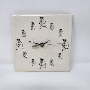 May include: Square ceramic clock with a cream-colored background and black pig illustrations. The clock face features black hands and a silver center. The pig illustrations are arranged around the clock face.