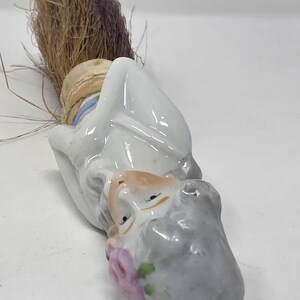 May include: A vintage porcelain figurine of a man with a grey beard and a brown straw broom. The figurine has a white coat, a tan face, and a pink flower. The broom is tied with a beige ribbon.