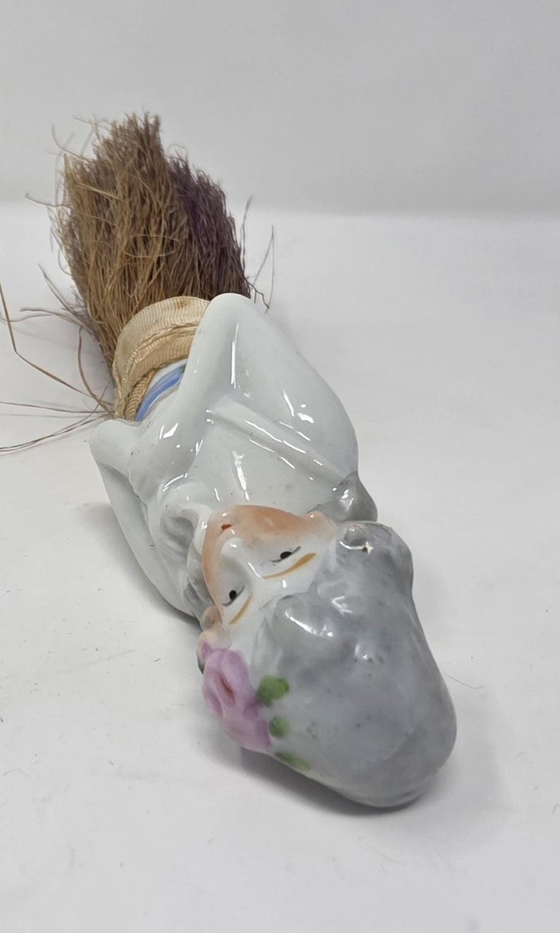 May include: A vintage porcelain figurine of a man with a grey beard and a brown straw broom. The figurine has a white coat, a tan face, and a pink flower. The broom is tied with a beige ribbon.