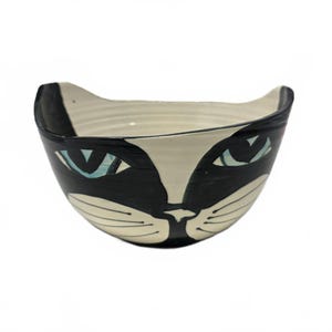 May include: A ceramic bowl shaped like a cat head with a black and cream color scheme. The design features pointed ears, light blue almond-shaped eyes, a small nose, and whiskers painted in black lines on a cream background.