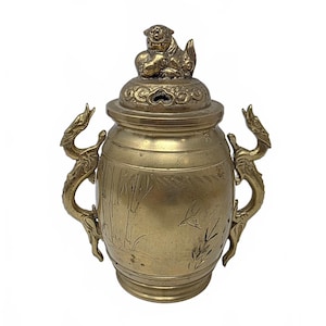 May include: A brass vessel with a lid featuring a sculpted lion finial. The body has two dragon-shaped handles on the sides and is decorated with etched bamboo and bird motifs. The metal has a muted gold tone with a smooth, polished finish.