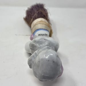 May include: A small, vintage porcelain clown figurine with a purple brush. The clown has a grey body, pink accents, and a blue and white collar. The brush is wrapped with a tan ribbon.