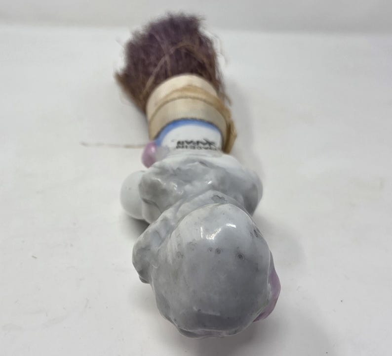 May include: A small, vintage porcelain clown figurine with a purple brush. The clown has a grey body, pink accents, and a blue and white collar. The brush is wrapped with a tan ribbon.