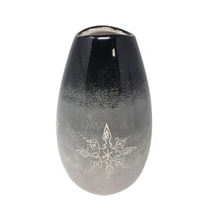 May include: A ceramic vase with a smooth, glossy finish featuring a gradient color transition from black at the top to a light gray at the base. The front of the vase is decorated with a white, etched snowflake design centered on the lower half.
