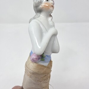 May include: A porcelain figurine of a woman with grey hair and a painted face. She is wearing a blue and pink floral sash and a beige fabric skirt. The figurine is attached to a brush with brown bristles.