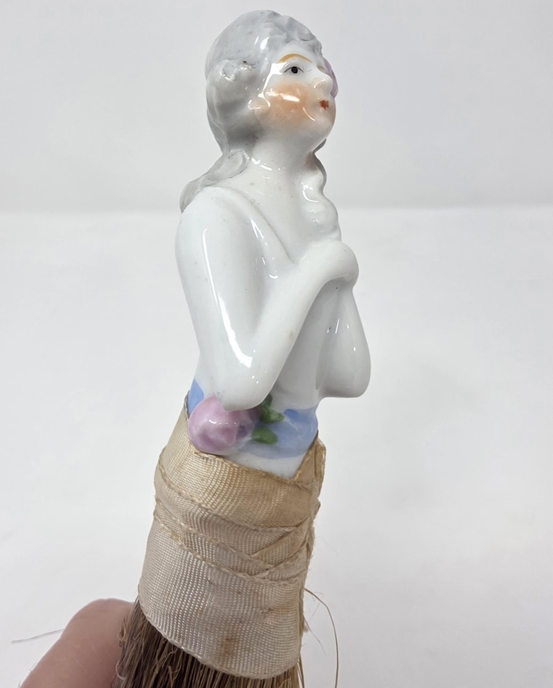 May include: A porcelain figurine of a woman with grey hair and a painted face. She is wearing a blue and pink floral sash and a beige fabric skirt. The figurine is attached to a brush with brown bristles.