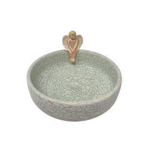May include: A round ceramic bowl featuring a light mint green glaze with a prominent dark crackle pattern throughout the surface. A small, light-colored figurine of an angel with wings is attached to the top rim, facing inward toward the center.