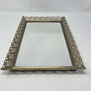 May include: A vintage vanity tray with a rectangular mirror. The tray has a decorative, filigree-style metal frame in a gold tone. The frame's edges are angled, and the mirror is clear.
