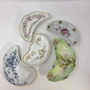 May include: Five antique, kidney-shaped porcelain dishes with floral designs. The dishes feature pink, purple, blue, and yellow flowers, with gold trim. The dishes are arranged on a white background.