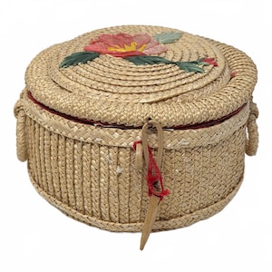 May include: A round, woven straw basket with a lid featuring a painted red flower with green leaves. The basket has a red fabric lining visible at the rim, two side handles, and a wooden latch closure secured with a thin red string.
