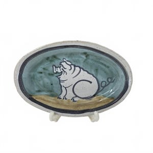 May include: Oval ceramic dish with a hand-painted pig illustration. The dish features a light blue interior with a black border and a beige base. The pig is white with black outlines and a curly tail. The dish is supported by a clear stand.