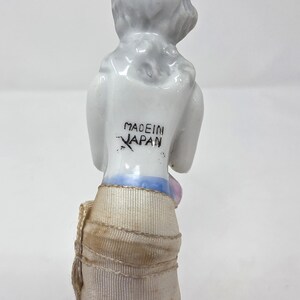 May include: A vintage porcelain figurine of a woman with a white head and shoulders, and a tan skirt. The words "MADE IN JAPAN" are printed on the back of the figurine. The figurine has a brush attached to the bottom.