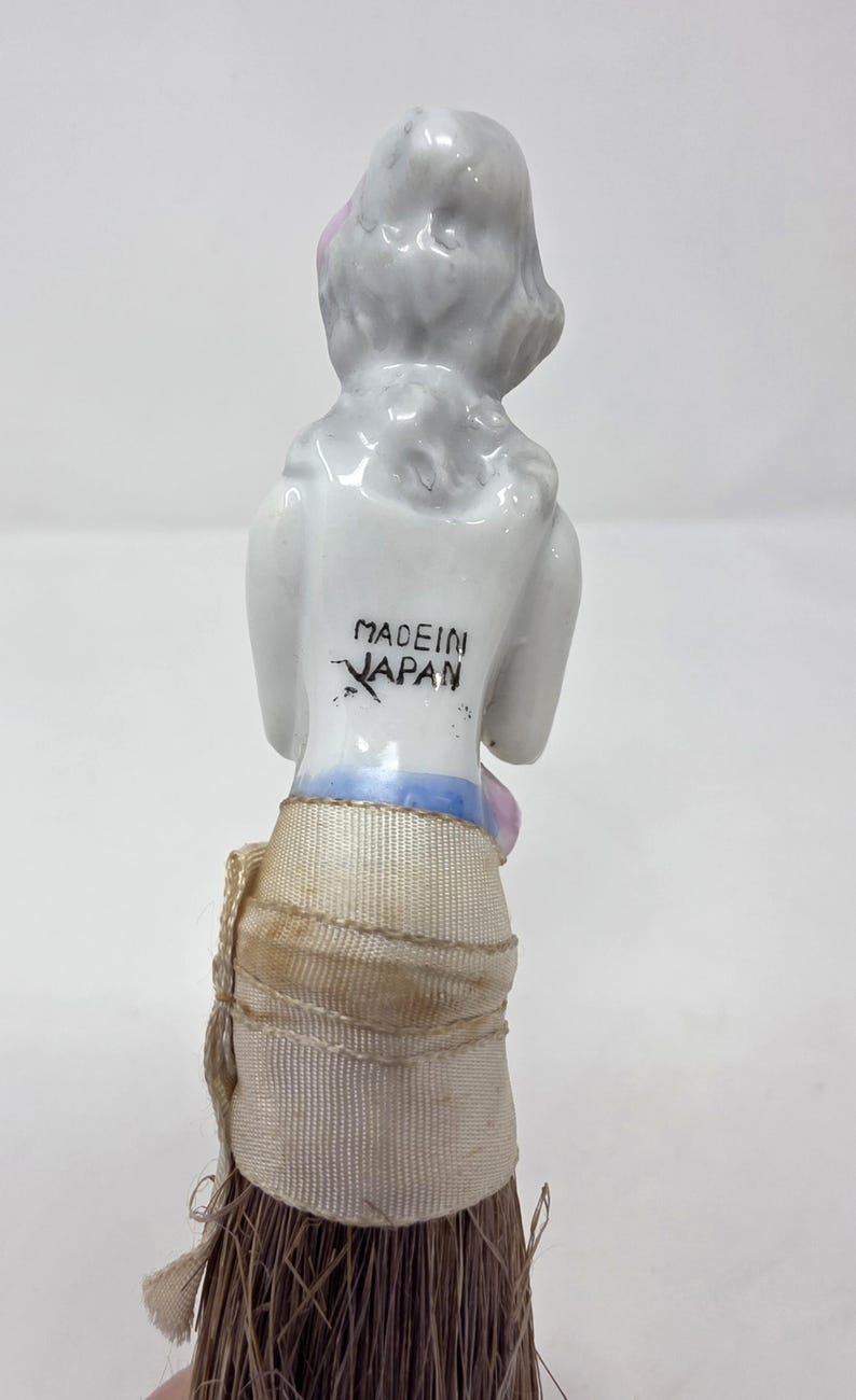 May include: A vintage porcelain figurine of a woman with a white head and shoulders, and a tan skirt. The words "MADE IN JAPAN" are printed on the back of the figurine. The figurine has a brush attached to the bottom.