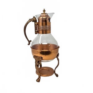 May include: A glass carafe with a copper-colored base and neck, featuring a decorative metal handle and a matching metal stand with three legs. The upper neck section has a perforated metal band, and the lid is topped with a small finial.