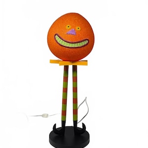 May include: A decorative lamp featuring a round, glittery orange head with yellow eyes, a purple nose, and a wide green grin. The head rests on a yellow star-shaped collar above two long, striped green and orange legs with black curled-toe shoes.