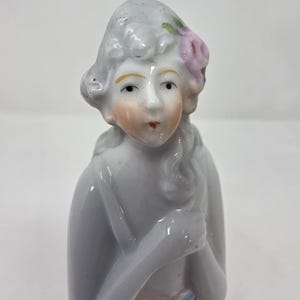 May include: A porcelain figurine of a woman with grey hair and a pink flower. The figurine has a pale complexion, rosy cheeks, and red lips. The woman is wearing a blue and gold garment. The figurine is a collectible item.