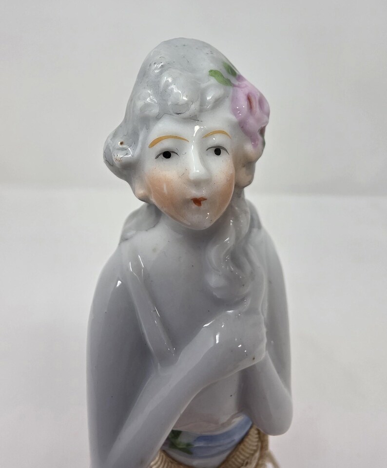 May include: A porcelain figurine of a woman with grey hair and a pink flower. The figurine has a pale complexion, rosy cheeks, and red lips. The woman is wearing a blue and gold garment. The figurine is a collectible item.