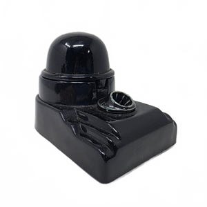 May include: A glossy black inkwell featuring a rounded dome-shaped lid and a rectangular base with a built-in pen rest. The inkwell has a smooth, reflective surface and a stepped design, with a small circular opening for a pen on the front section.