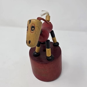 May include: A wooden pull-along toy horse with a red body, tan head, and black and tan legs. The horse has painted details and a red string tail. The toy is mounted on a red cylindrical base.