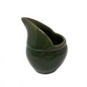 May include: A small, olive green ceramic creamer pitcher with a glossy finish. The vessel features a rounded, bulbous body and a high, elongated pouring spout that curves upward, creating a distinct asymmetrical silhouette with a wide opening.