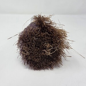 May include: A close-up shot of a brown, spherical brush head with a clear handle. The brush head is made up of many thin, bristly fibres, with some stray fibres extending outwards. The brush is set against a plain white background.