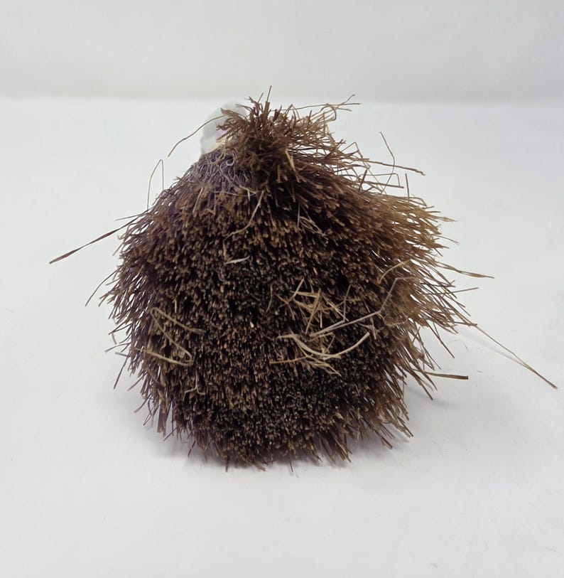 May include: A close-up shot of a brown, spherical brush head with a clear handle. The brush head is made up of many thin, bristly fibres, with some stray fibres extending outwards. The brush is set against a plain white background.